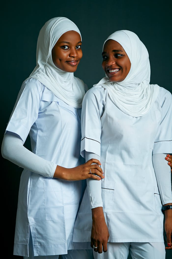 Two female nurses wearing hijabs and white uniforms, smiling warmly.